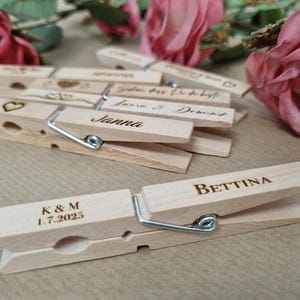 Puede incluir: Pinzas de madera claras grabadas con nombres y fechas, ideales para recuerdos de boda o marcadores de sitio. Las pinzas tienen muelles de metal plateado. Rosas rosas en el fondo. Algunas pinzas tienen un símbolo de corazón.
