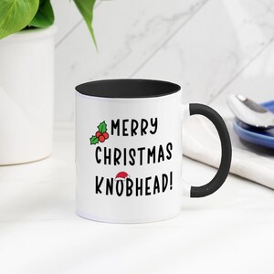 May include: White ceramic coffee mug with a black interior and handle. The mug has a black and green holly sprig with red berries and the text "Merry Christmas Knobhead!" with a Santa hat on the word "Knobhead".