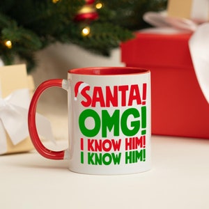 May include: White ceramic mug with a red rim and handle. The mug has a green and red design with the text "SANTA! OMG! I KNOW HIM! I KNOW HIM!"