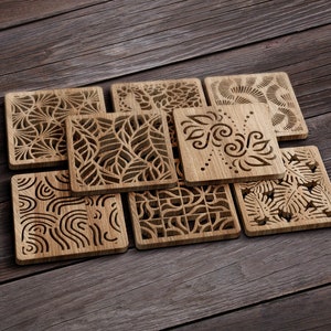 Puede incluir: Ocho posavasos de madera con intrincados diseños cortados con láser. Cada posavasos presenta un patrón diferente, que incluye formas geométricas, hojas y espirales. Los posavasos están dispuestos en un patrón escalonado sobre una superficie de madera.