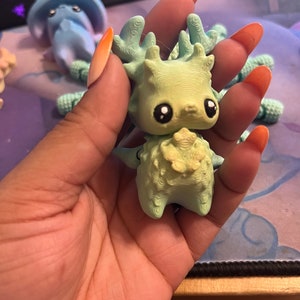 May include: A light green figurine of a creature with large eyes, antlers, and a textured body. The figurine is held in a hand, with other similar figurines visible in the background. The figurine has a whimsical, fantasy theme.