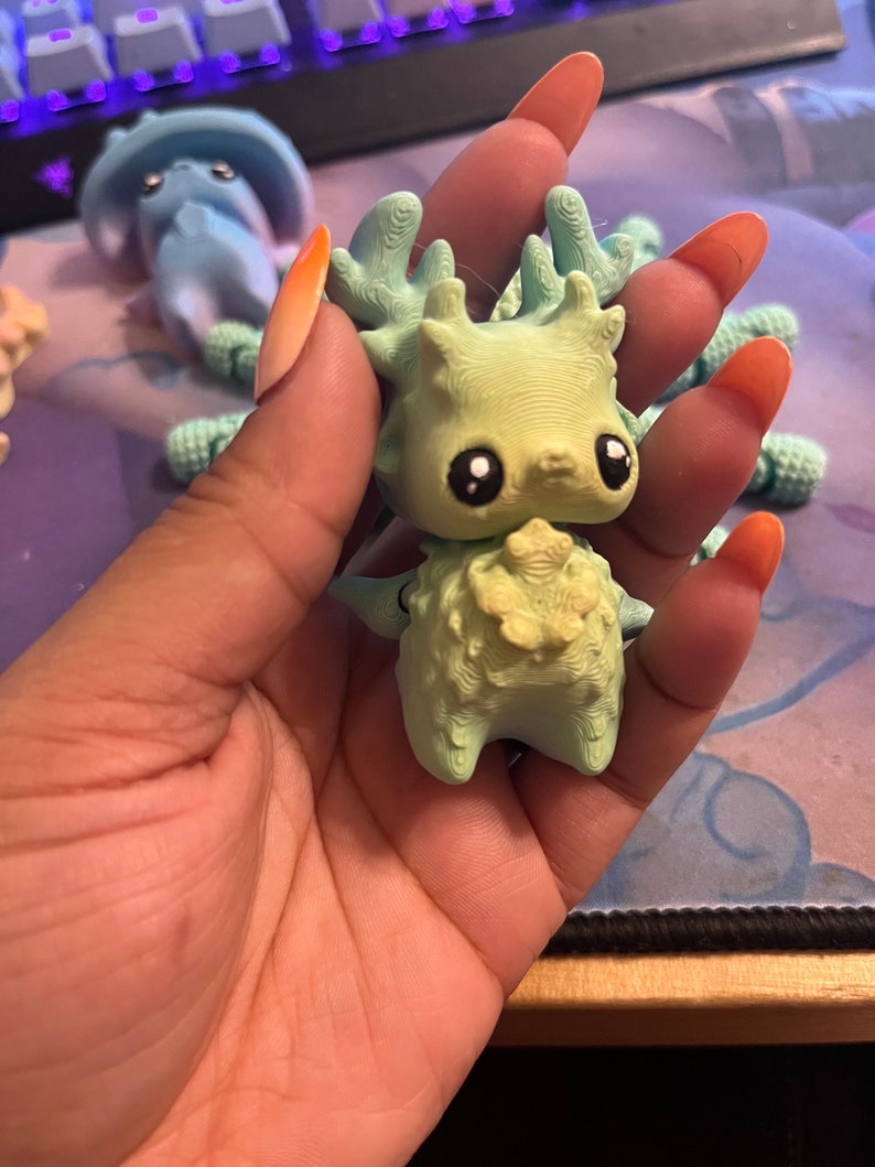 May include: A light green figurine of a creature with large eyes, antlers, and a textured body. The figurine is held in a hand, with other similar figurines visible in the background. The figurine has a whimsical, fantasy theme.