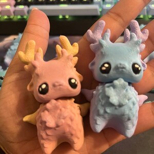 May include: Two small, whimsical figurines in a hand. One is pink with yellow antlers, the other is light blue with purple antlers. Both have large black eyes and small arms.