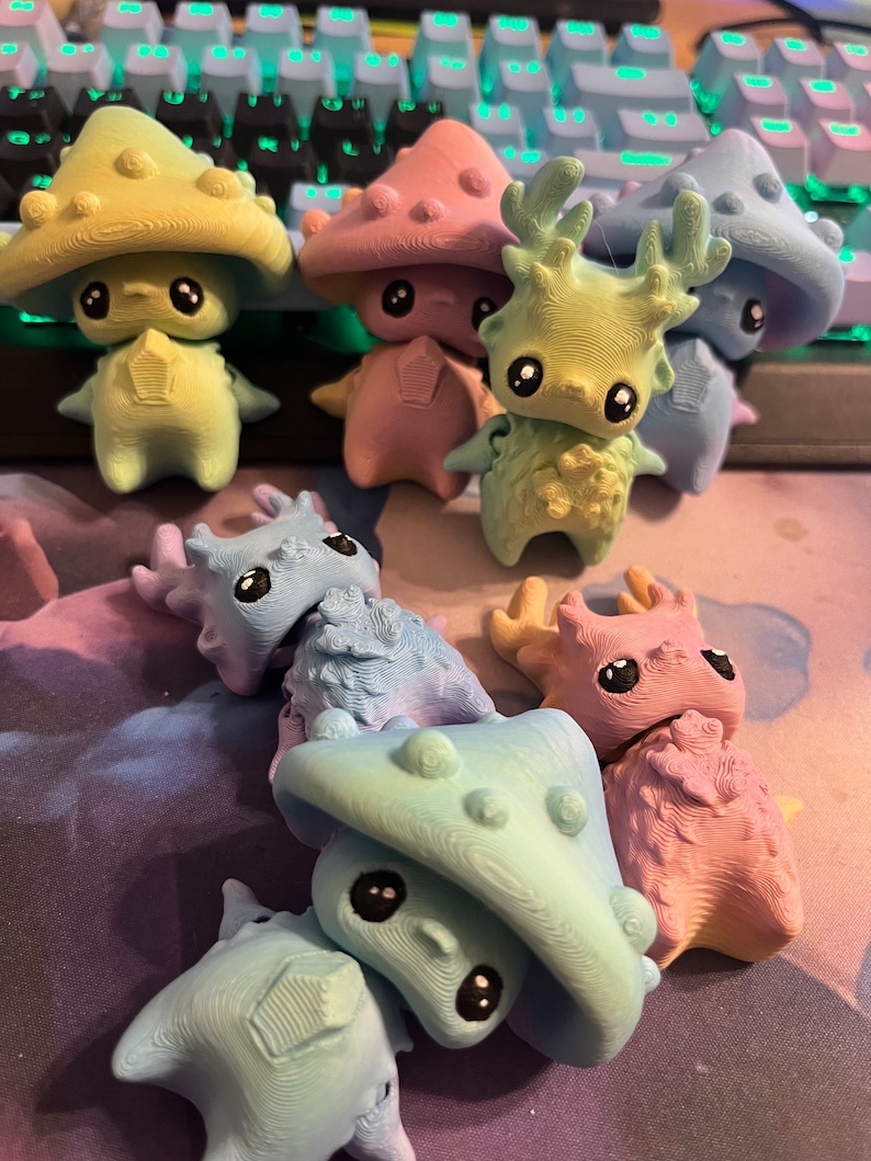 May include: A collection of colorful, 3D-printed figurines. The figures include mushroom-shaped characters in pastel yellow, pink, and blue, and a green character with antlers. Each figure has large, black eyes and a whimsical design.