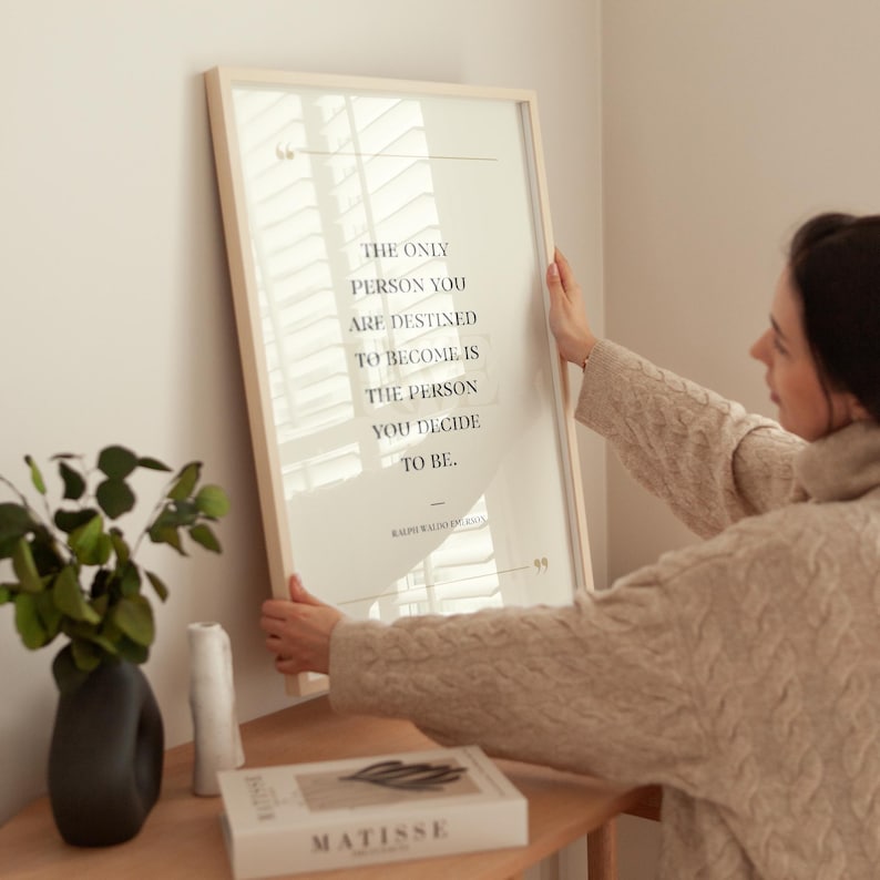K&ouml;nnte beinhalten: Ein gerahmter Druck mit dem Zitat &bdquo;THE ONLY PERSON YOU ARE DESTINED TO BECOME IS THE PERSON YOU DECIDE TO BE.&ldquo; Der Druck befindet sich in einem hellen Rahmen. Eine Person h&auml;lt den Rahmen. Eine Vase und ein Buch liegen auf einer Holzoberfl&auml;che.