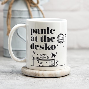 May include: White ceramic mug with a black design that says "panic at the desko" with a disco ball and a cat at a desk.
