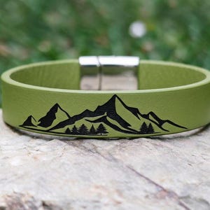 Op de afbeelding: Een saliegroene leren armband met een zwarte grafische print van een bergketen met dennenbomen aan de voet. De armband is voorzien van een gepolijste zilverkleurige magnetische metalen sluiting, wat zorgt voor een gladde, ronde band.