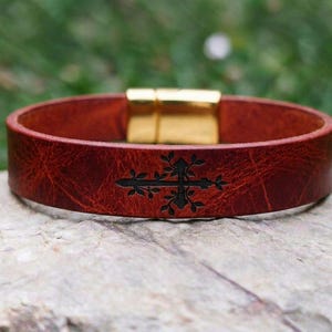 Op de afbeelding: Een brede, roodbruine leren armband met een donker, gegraveerd bloemmotief in de vorm van een kruis in het midden. De armband is voorzien van een gepolijste, goudkleurige magnetische metalen sluiting voor een veilige bevestiging.
