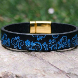 Op de afbeelding: Een zwarte leren armband met een levendig blauw bloemen- en krulpatroon dat in de band is gedrukt. De armband wordt vastgezet met een gepolijste goudkleurige magnetische metalen sluiting, wat zorgt voor een gladde, ronde vorm.