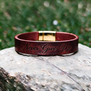 Op de afbeelding: Een donkerrode leren armband met de gegraveerde tekst "Never Give Up" in een cursief lettertype. De armband is voorzien van een gepolijste goudkleurige metalen magnetische sluiting, wat zorgt voor een strakke afwerking van het ontwerp.