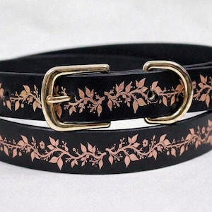 Op de afbeelding: Een zwarte riem met een doorlopend koperkleurig bloemenrankpatroon dat over de lengte van de band is aangebracht. De riem is voorzien van een gepolijste goudkleurige metalen gesp en een bijpassende goudkleurige D-ringlus voor de riemuiteinde.