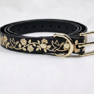 Op de afbeelding: Een zwarte riem met een goudkleurig bloemen- en vogelpatroon langs de band. De riem is voorzien van een goudkleurige metalen gesp en een bijpassende goudkleurige D-ring, met een reeks verstelgaatjes langs de lengte van de zwarte band.
