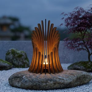 Op de afbeelding: Een bruine houten kaarsenhouder met een reeks verticale, gebogen latten die in een waaiervorm zijn gerangschikt. Een klein, brandend waxinelichtje staat in het midden en werpt een warm, gloeiend licht door de houten latten heen.
