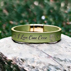 Op de afbeelding: Een licht olijfgroene leren armband met een goudkleurige magnetische metalen sluiting. Het oppervlak is gegraveerd met de tekst "I Love Cane Corso", geflankeerd door twee kleine hartjes en een gedetailleerde lijntekening van een Cane Corso.