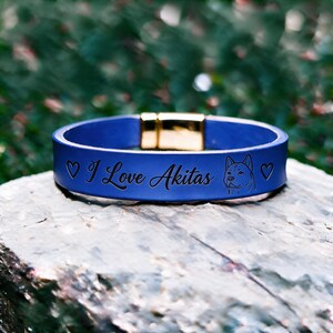 Könnte beinhalten: Ein blaues Lederarmband mit einem goldfarbenen Metallverschluss. Die Oberfläche ist mit dem Text I Love Akitas graviert, flankiert von zwei kleinen Herzsymbolen und einer Strichzeichnung eines Akita-Hundekopfes auf der rechten Seite.