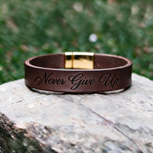 Op de afbeelding: Een bruine leren armband met de gegraveerde tekst Never Give Up in een cursief lettertype. De armband is bevestigd met een gepolijste goudkleurige metalen magnetische sluiting, wat een gladde afwerking geeft aan het ontwerp van het accessoire.