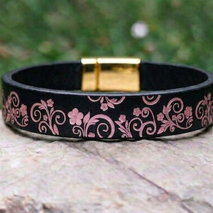 Op de afbeelding: Een zwarte leren armband met een herhalend roze bloemen- en krulpatroon dat in de band is gegraveerd. De armband wordt vastgezet met een rechthoekige goudkleurige metalen magnetische sluiting die de uiteinden van de band verbindt.