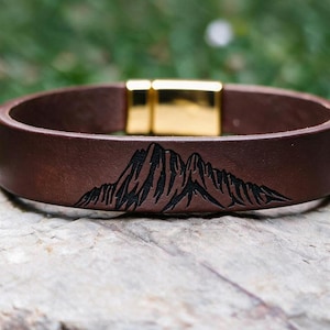 Op de afbeelding: Een bruine leren armband met een zwarte gravure van een bergketen. De armband is voorzien van een gepolijste goudkleurige magnetische metalen sluiting, wat zorgt voor een gladde en strakke afwerking van het uiterlijk van de band.