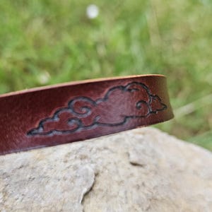 Op de afbeelding: Close-up van een donkerbruine leren armband met een wolkenontwerp. Het wolkenontwerp is in het leer geëtst en heeft een donkerdere bruine tint. De armband ligt op een steen.