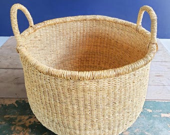 May include: A large, round, natural woven basket with two handles. The basket is made of a light brown, natural material and has a smooth, even texture.
