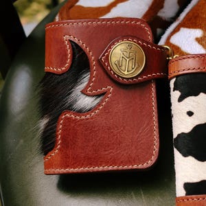 May include: A brown leather wallet with a decorative black and white cowhide accent. The wallet features a brass clasp with an anchor emblem and white stitching. The design is rustic and handcrafted.