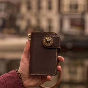 Op de afbeelding: Een bruine leren portemonnee met een messing embleem met een schedelontwerp. De portemonnee heeft een gestikte rand en een lus voor een ketting. De achtergrond is wazig en toont een gebouw.