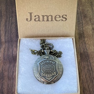 May include: A gold-toned pocket watch with a chain, featuring a police badge design with the words "To Protect and Serve" and "Police" inscribed on it. The watch is in a brown box with the name "James" engraved on the top.