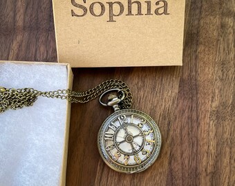 May include: A gold-toned pocket watch necklace with a white face and black Roman numeral markings. The watch is on a chain and comes in a brown box with the name "Sophia" engraved on the top.