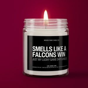 May include: A white candle in a clear glass jar with a black label that reads "Wicked Funny Candle Co. Smells Like A Falcons Win Just My Lucky Game Day Candle". The candle is lit and the flame is visible.