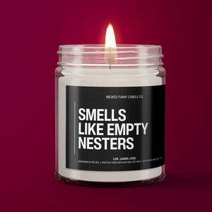 May include: A white candle in a clear glass jar with a black label that reads "WICKED FUNNY CANDLE CO. SMELLS LIKE EMPTY NESTERS LIVE. LAUGH. LOVE. HANDMADE IN THE USA | CRAFTED FROM 100% NATURAL SOY WAX"