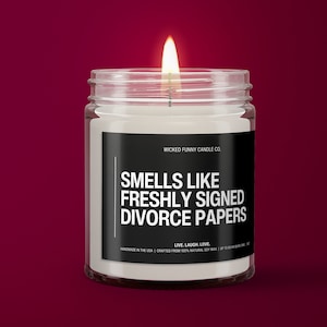 May include: A white candle in a clear glass jar with a black label that reads "WICKED FUNNY CANDLE CO. SMELLS LIKE FRESHLY SIGNED DIVORCE PAPERS LIVE. LAUGH. LOVE. HANDMADE IN THE USA | CRAFTED FROM 100% NATURAL SOY WAX | UP TO 60 HOUR BURN TIME | 10 OZ"