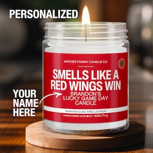 May include: Personalized candle with a red and white label that says "Smells Like a Red Wings Win" and "Brandon's Lucky Game Day Candle". The candle is in a clear glass jar and is lit.