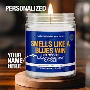 May include: A white candle in a clear glass jar with a blue and yellow label that reads "Smells Like A Blues Win Brandon's Lucky Game Day Candle" and "Winning Sure Smells Good". The label also includes a small heart and the text "Hand Poured With By: Wicked Funny".