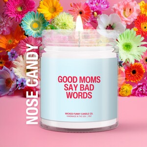 May include: A lit candle in a glass jar with a light blue label that reads "GOOD MOMS SAY BAD WORDS" in red text. The candle is surrounded by colorful flowers against a pink background. The text "NOSE CANDY" is printed vertically on the left side.