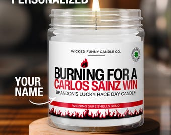 May include: A lit, clear glass candle jar with a white label. The label reads "BURNING FOR A CARLOS SAINZ WIN" with the text "BRANDON'S LUCKY RACE DAY CANDLE". The candle sits on a small wooden coaster. The text "WINNING SURE SMELLS GOOD" is also visible.