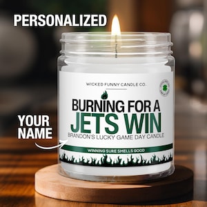 May include: A lit, clear glass candle jar with a white label that reads "BURNING FOR A JETS WIN". The candle is on a wooden coaster. The text "WICKED FUNNY CANDLE CO." is above the main text. The words "YOUR NAME" and "PERSONALIZED" are also visible.