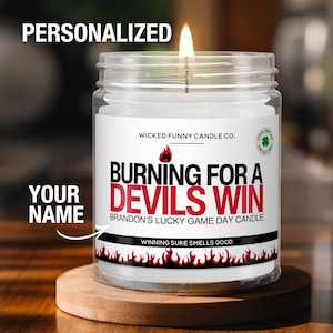 May include: A lit glass candle with a white label that reads "BURNING FOR A DEVILS WIN" in red and black text. The candle is personalized with the text "YOUR NAME" and sits on a wooden coaster. The candle is from Wicked Funny Candle Co.