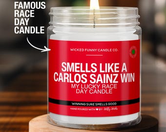 May include: A red and white candle with the text "Smells Like A Carlos Sainz Win My Lucky Race Day Candle" and "Wicked Funny Candle Co.". The candle is on a wooden surface.