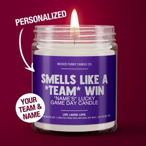 May include: A lit, clear glass jar candle with a purple label that reads "Smells Like a *Team* Win". The candle is personalized with the text "Your Team & Name". The candle is made in the USA with natural soy wax.