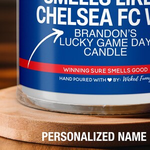 May include: A blue and red candle with the text "CHELSEA FC" and "BRANDON'S LUCKY GAME DAY CANDLE". The text "WINNING SURE SMELLS GOOD" is on a red stripe. The candle is on a wooden surface.