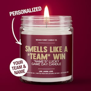 May include: A personalized candle with a burgundy label that reads "Smells Like A *Team* Win *Name's* Lucky Game Day Candle". The candle is in a clear glass jar with a white wax interior and a lit wick. The label also includes the text "Wicked Funny Candle Co.", "Live. Laugh. Love.", "Made in the USA", "Crafted from 100% Natural Soy Wax", and "60 HR Burn Time".