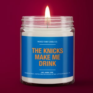 May include: A glass candle jar with a blue label that reads "WICKED FUNNY CANDLE CO. THE KNICKS MAKE ME DRINK LIVE. LAUGH. LOVE. HANDMADE IN THE USA | CRAFTED FROM 100% NATURAL SOY WAX | UP TO 60 HR BURN TIME | 8OZ".