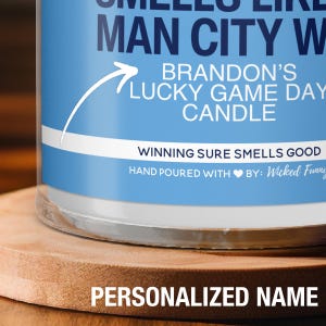 Puede incluir: Una vela azul y blanca con el texto "Brandon's Lucky Game Day Candle" y "Winning Sure Smells Good". La vela est&aacute; hecha a mano con un s&iacute;mbolo de coraz&oacute;n y el texto "By: Wicked Funny".