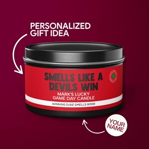 May include: A red and black candle tin with the text "Smells Like A Devils Win" and "Mark's Lucky Game Day Candle". The text "Winning Sure Smells Good" is printed below. The text "Your Name" is printed on a white circle below the candle tin.