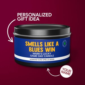May include: A black tin candle with a blue and yellow label that reads "Smells Like A Blues Win Mark's Lucky Game Day Candle Winning Sure Smells Good". The label also has a small green shamrock logo. The candle is a personalised gift idea.