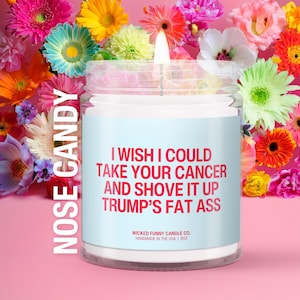 May include: A clear glass jar candle with a light blue label featuring red text that reads: NOSE CANDY, I WISH I COULD TAKE YOUR CANCER AND SHOVE IT UP TRUMP'S FAT ASS, WICKED FUNNY CANDLE CO. HANDMADE IN THE USA | 9OZ. The candle has a lit wick.