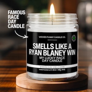 May include: A black and white candle with the text "Smells Like A Ryan Blaney Win" and "My Lucky Race Day Candle". The candle is in a clear glass jar with a black label and a green shamrock logo.