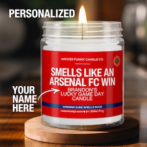 May include: A red and white candle with the text "Smells Like An Arsenal FC Win" and "Brandon's Lucky Game Day Candle". The candle is in a clear glass jar with a red lid and is sitting on a wooden surface.