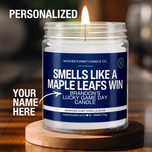 May include: A clear glass candle jar with a white candle inside. The label is blue and white and says "Smells Like A Maple Leafs Win" and "Brandon's Lucky Game Day Candle".