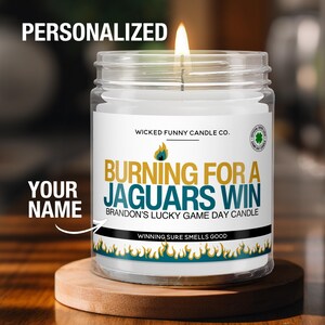 May include: A lit, personalized candle in a clear glass jar. The label reads "Burning for a Jaguars Win" with the text "Your Name" and "Brandon's Lucky Game Day Candle." The candle sits on a wooden coaster.